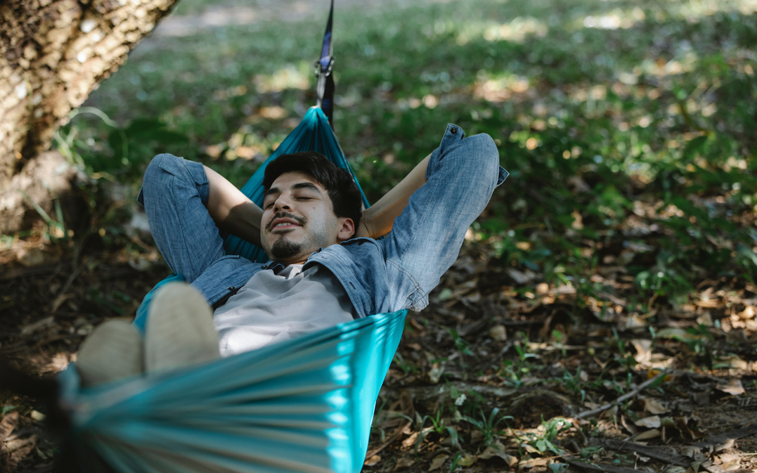 Young man relaxing in teal hammock under trees in a sun dappled park