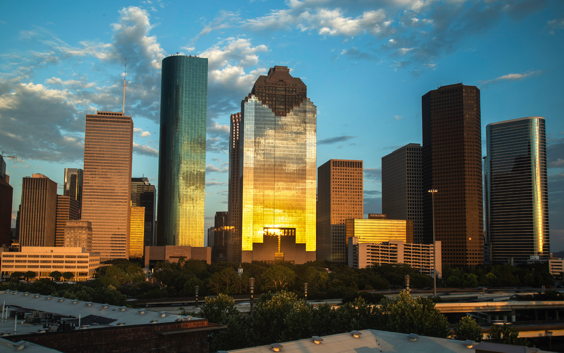 Downtown Houston skyline at golden hour with sunlight reflecting off glass skyscrapers and trees in the foreground