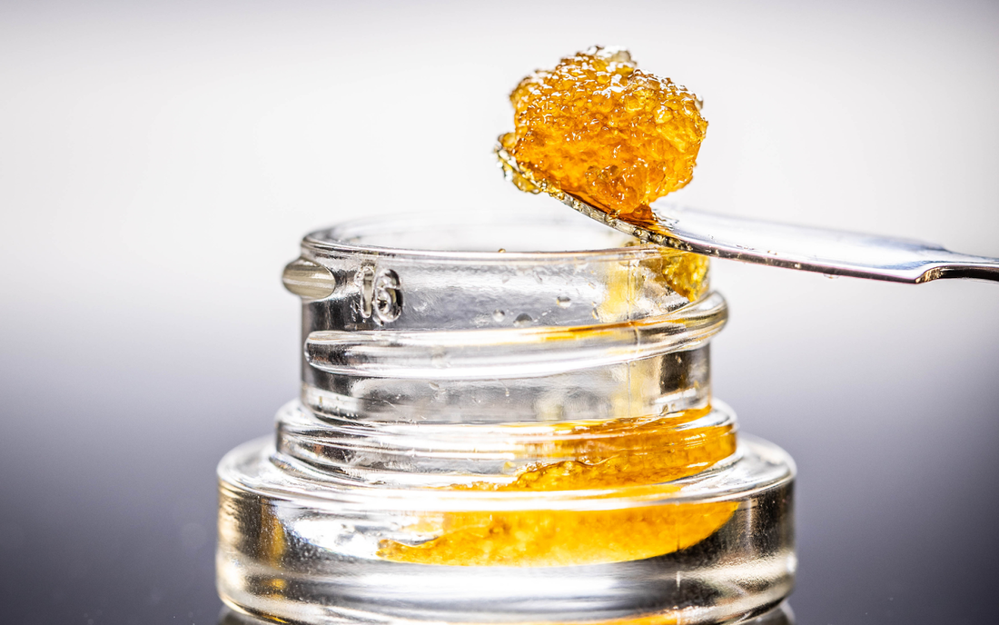Jar of amber cannabis concentrate with a metal dab tool lifting a sticky glob against a pale gray background