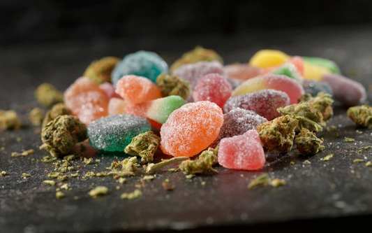 Assorted sugar-coated THC gummies scattered with cannabis buds on a dark slate surface