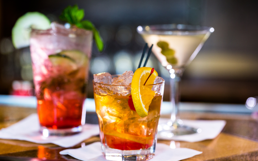 Three cocktails on a bar top with citrus and cherry garnishes and a blurred bar background