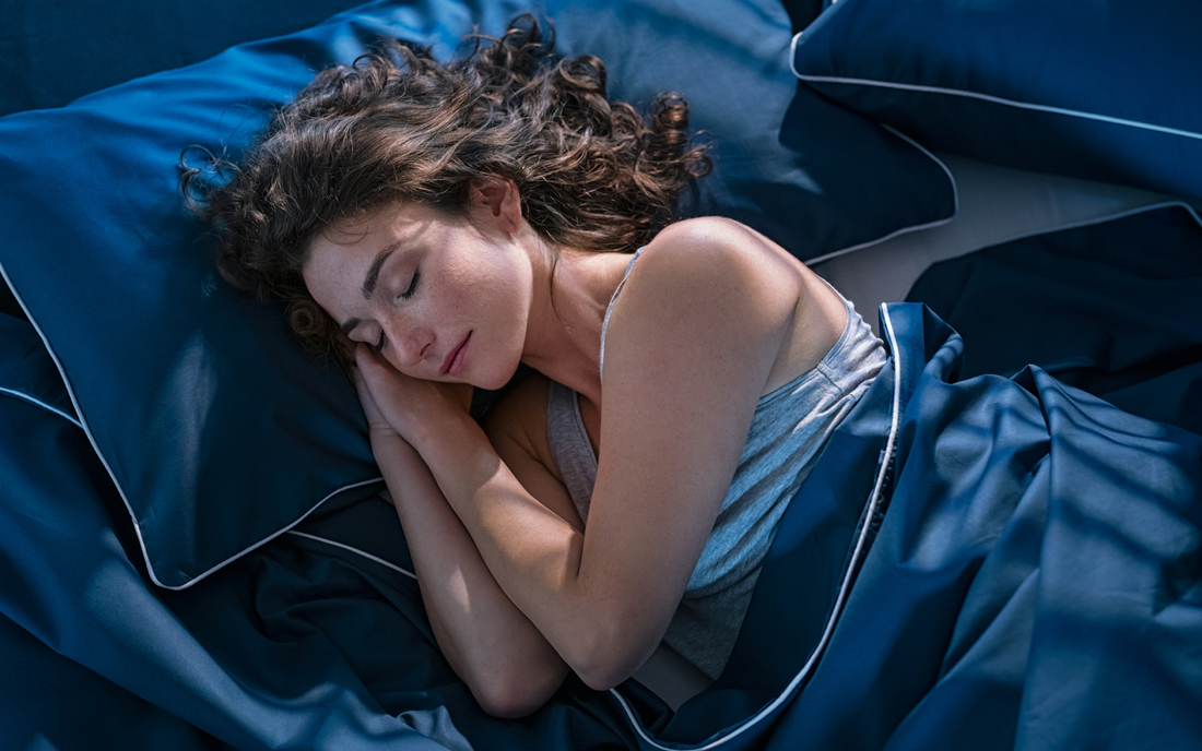 Woman sleeping on her side in bed under navy blue sheets and pillows, appearing relaxed