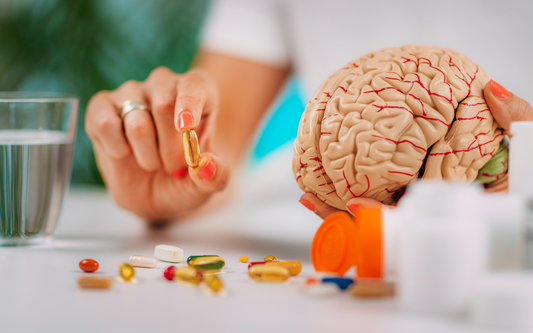 Hands holding a brain model and picking up a golden supplement capsule from a table scattered with pills and an open orange pill bottle