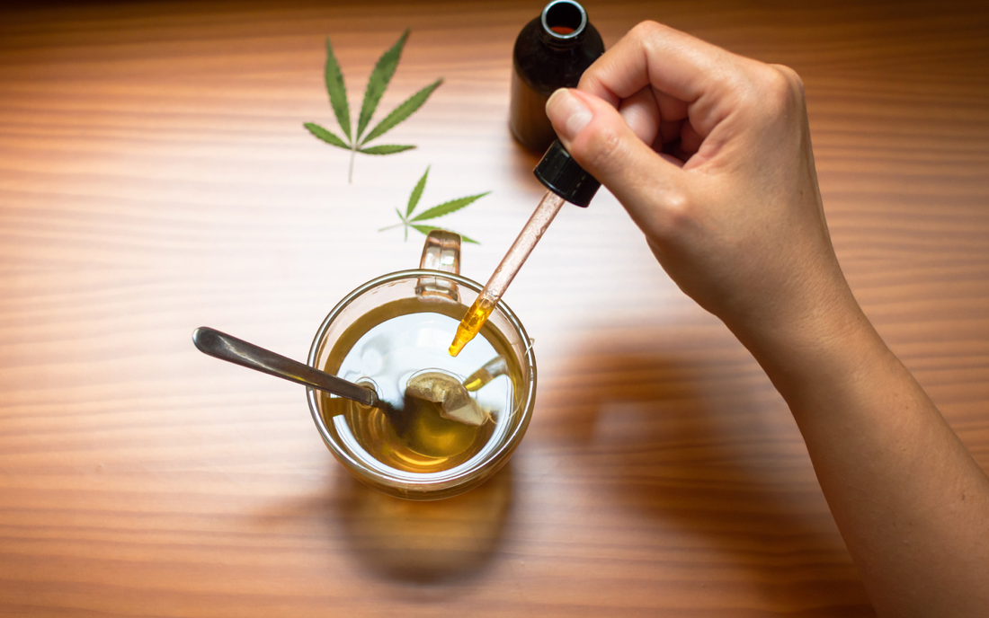 Hand using dropper to add cannabis tincture to a cup of tea on a wooden table with cannabis leaves nearby