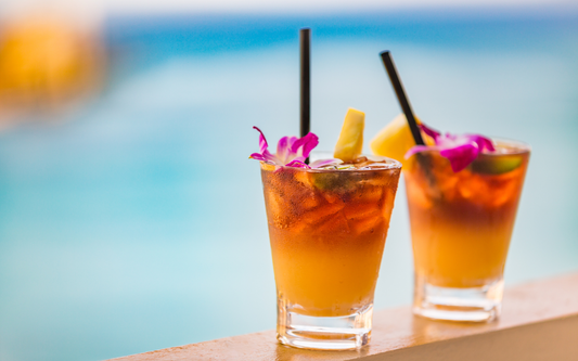 Two tropical mocktails on a ledge by a pool garnished with pineapple wedges and purple orchid flowers