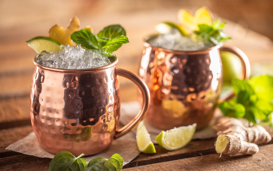 Two copper Moscow Mule mugs with crushed ice, lime wedges, and mint on a wooden table with ginger root