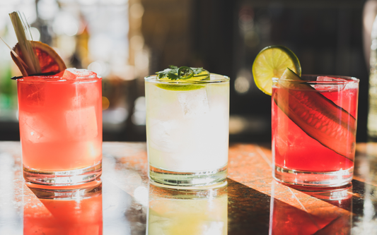 Three mocktails on a bar: two pink drinks and a pale lime mocktail garnished with lime slice and jalapeño, warm backlit setting