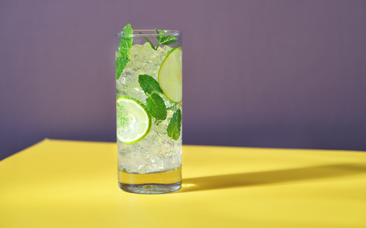 Glass of mojito mocktail with crushed ice, lime slices, and fresh mint on a yellow table against a purple background