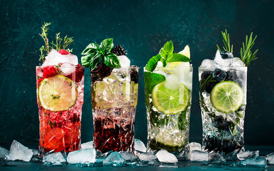 Four colorful mocktails in tall glasses garnished with lime slices, berries, and fresh herbs on crushed ice against a dark teal background