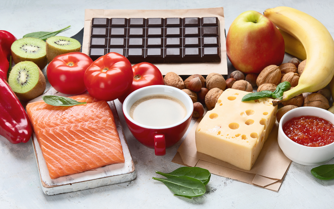 Serotonin-linked foods arranged on a table, including salmon, dark chocolate, Swiss cheese, nuts, banana, apple, kiwi, tomatoes, and a cup of coffee