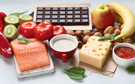 Serotonin-linked foods arranged on a table, including salmon, dark chocolate, Swiss cheese, nuts, banana, apple, kiwi, tomatoes, and a cup of coffee