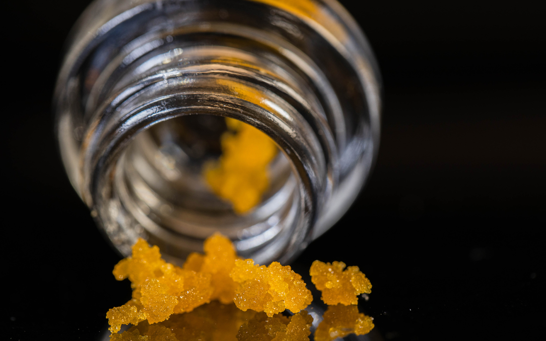 Close-up of golden Delta-9 THC concentrate crumbs spilling from a small glass jar on a black background