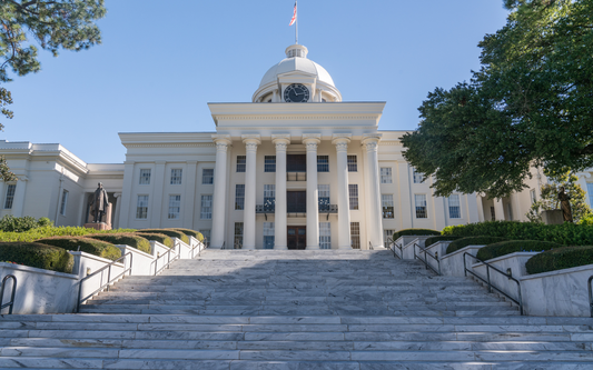 Front view of a state capitol building with marble steps, six white columns and a domed clock tower against a clear blue sky
