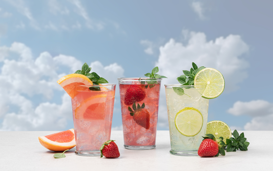 Three fruit-infused drinks garnished with grapefruit, strawberries, and lime on a white surface against a cloudy blue sky