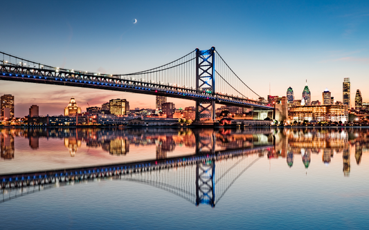 Suspension bridge at dusk spanning a river with a lit city skyline reflected in calm water