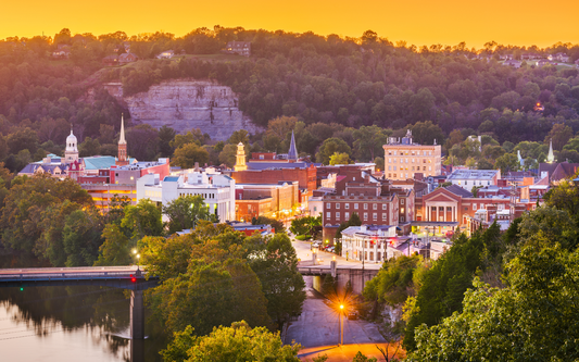 Sunset view of a Kentucky riverfront town with downtown buildings, a bridge over the river, and wooded cliffs behind