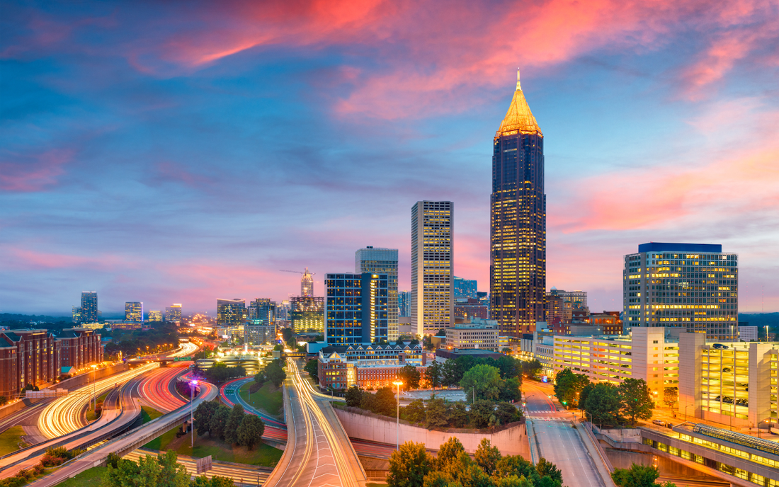 City skyline at dusk with illuminated skyscrapers and winding highway light trails under a pink-blue sky