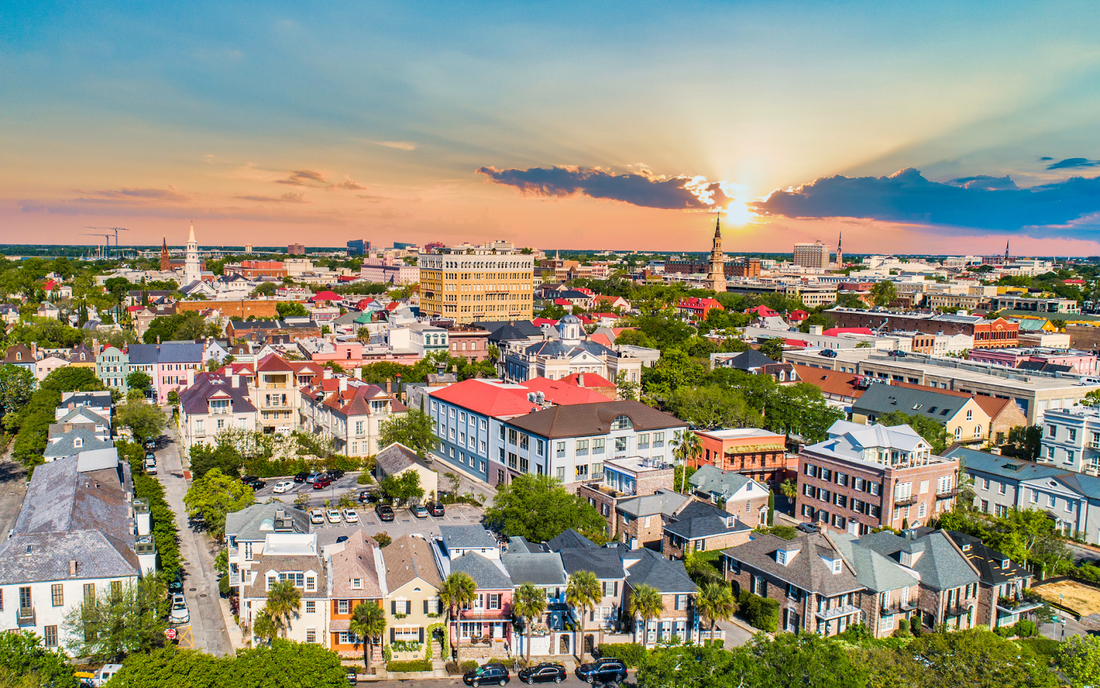 Aerial view of a coastal city at sunset with colorful historic homes, church spires, and sun rays breaking through clouds