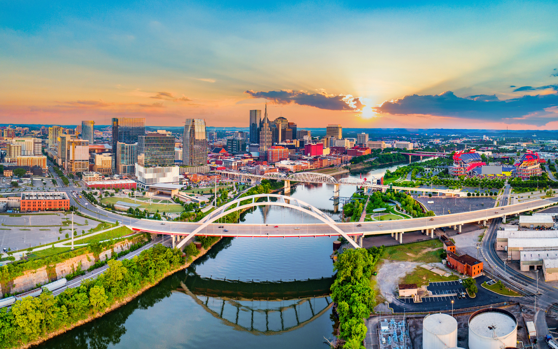 Nashville skyline at sunset with downtown high-rises, bridges crossing the Cumberland River, and a stadium visible to the right