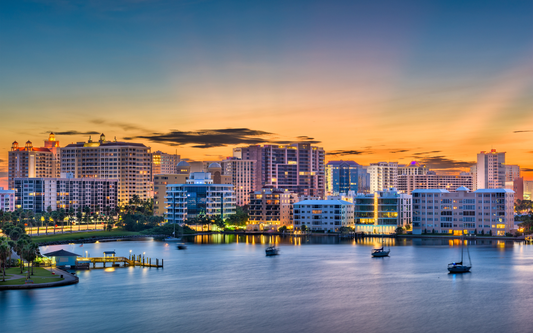 Waterfront Florida city skyline glowing at sunset with lit high-rise condos and sailboats on calm water