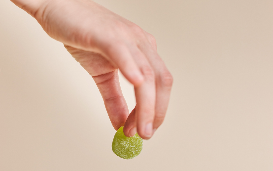 Hand holding a green sugar-coated CBD gummy between thumb and index finger against a beige background
