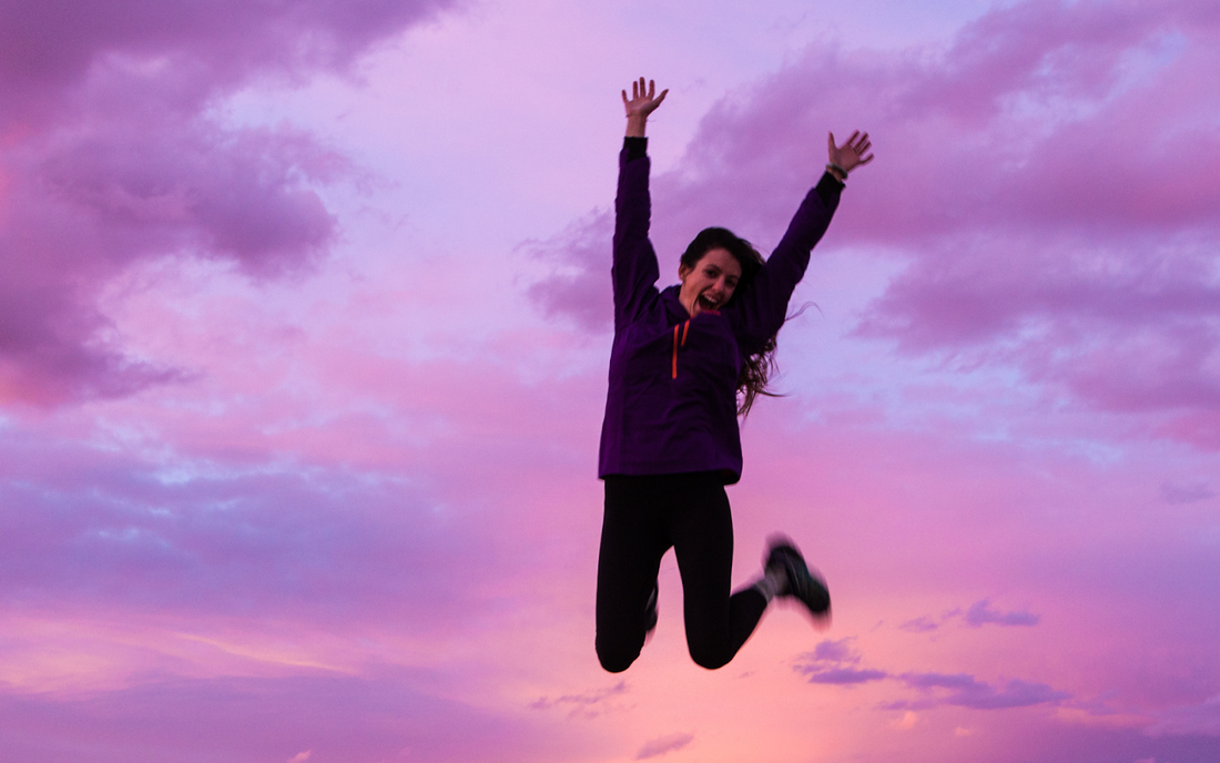 Woman jumping with arms raised against a pink and purple sunset sky