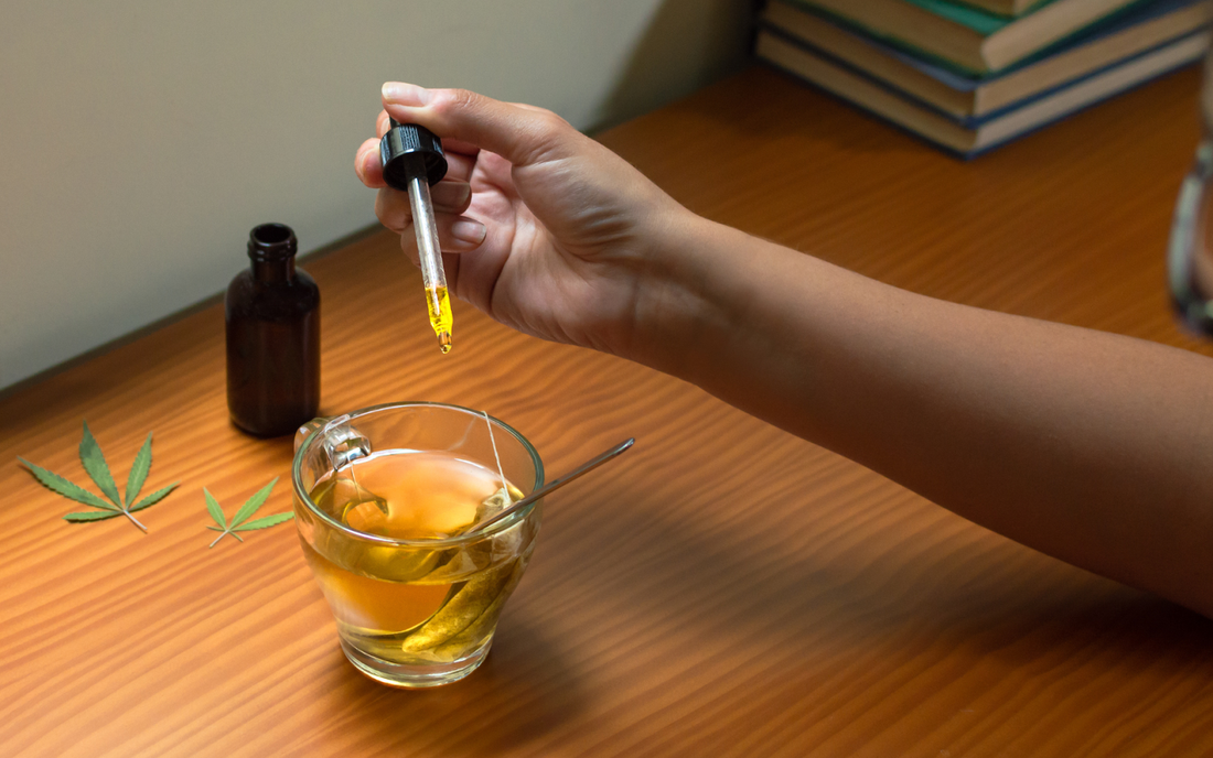 Hand dropping CBD oil from a dropper into a cup of tea on a wooden table with hemp leaves and an amber oil bottle