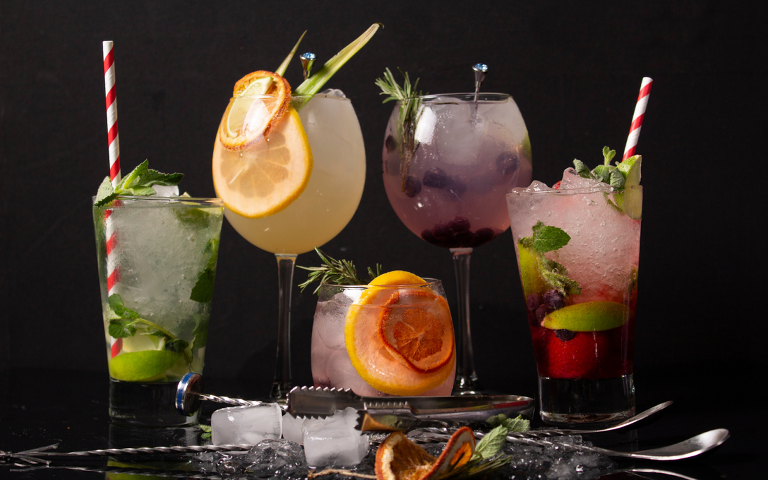 Five cocktails on a black tabletop, garnished with citrus slices, berries and mint, with ice and bar tools