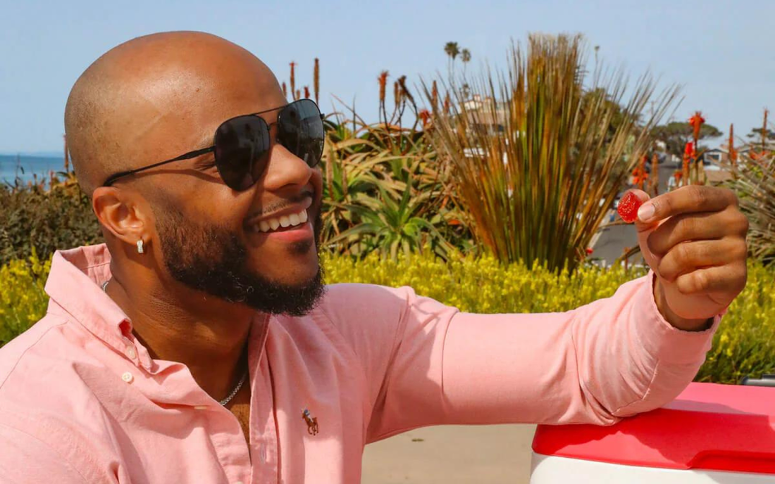 Smiling man in sunglasses holding a red CBD gummy at a sunny beachside park