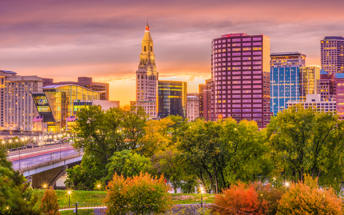 Hartford skyline glowing at sunset with downtown towers, a highway bridge, and a tree-lined park in the foreground