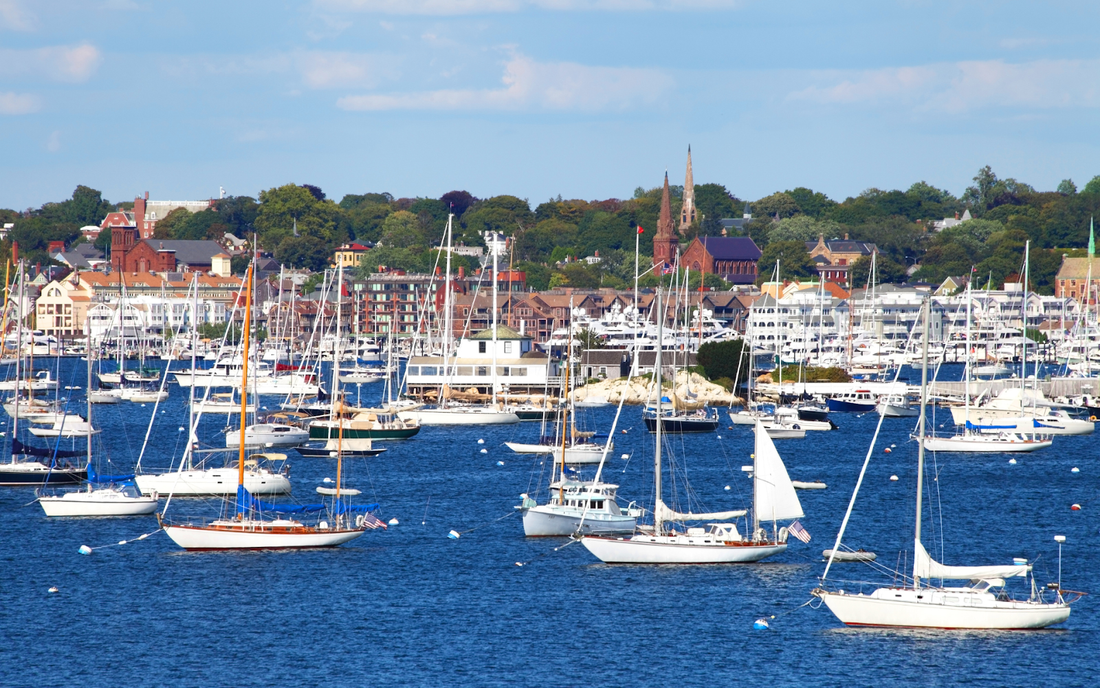 Sailboats moored in a busy harbor with a coastal town and church steeples in the background