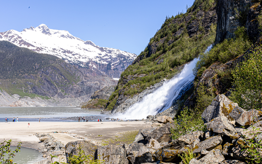 Visitors on a sandy shore beside a large waterfall cascading down steep, snow‑capped Alaskan mountains