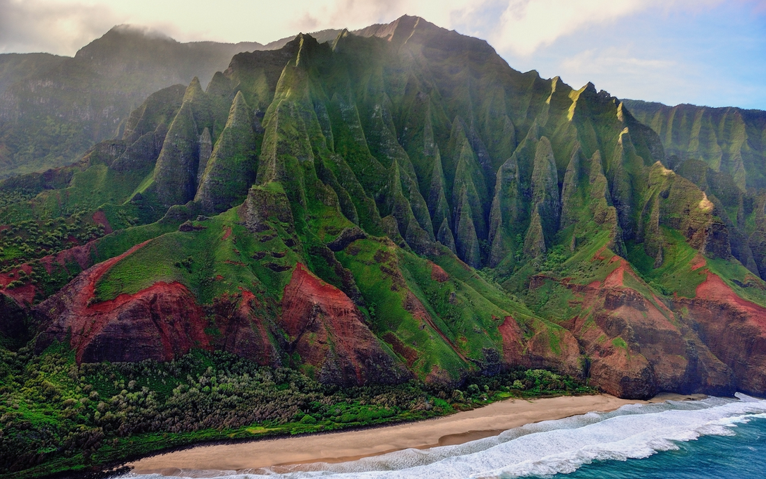 Aerial view of steep green cliffs and red earth along a Hawaiian coastline above a sandy beach and breaking surf