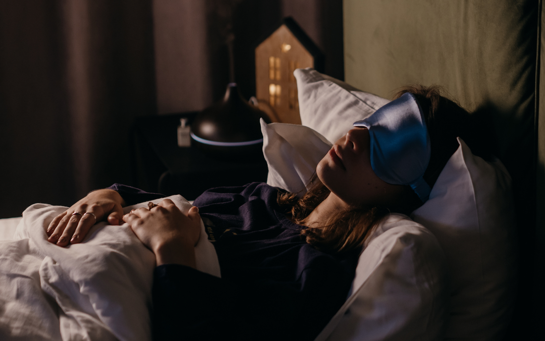 Person lying in bed wearing a blue sleep mask, hands on blanket in a dimly lit bedroom with a diffuser on the nightstand