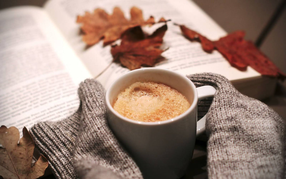 Cup of coffee resting on a gray knit sweater beside an open book with autumn leaves, warm cozy lighting