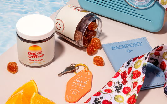 Jar of blood orange gummies spilling onto a pastel tabletop beside a blue passport, orange slice, and beach please keychain