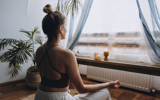 Woman sitting cross-legged meditating by a window with lit candles and a potted palm, soft natural light