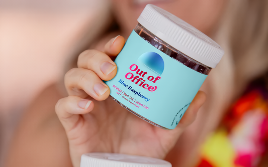 Close-up of jar of Out of Office Blue Raspberry THC gummies showing turquoise label reading Out of Office and white lid, held in hand against blurred background