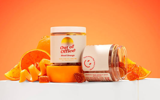 Front view of Out of Office Blood Orange gummy jars, one upright and one tipped showing sugar-coated gummies and sliced blood orange on orange background