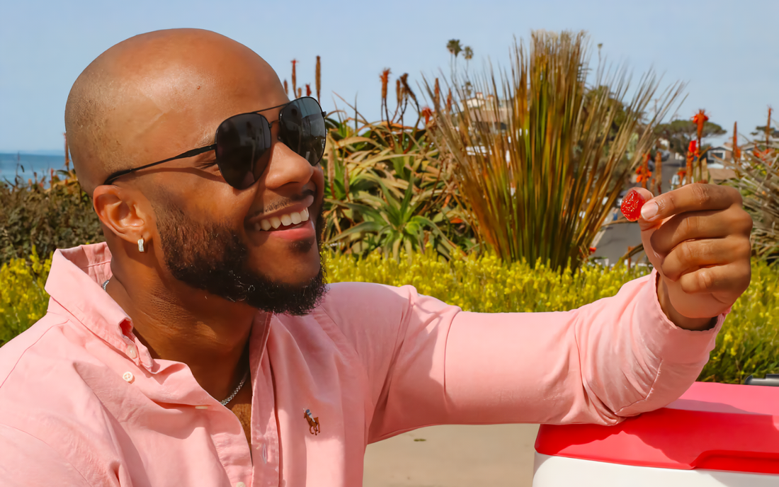 Smiling man in sunglasses holding a red gummy near coastal plants and the ocean