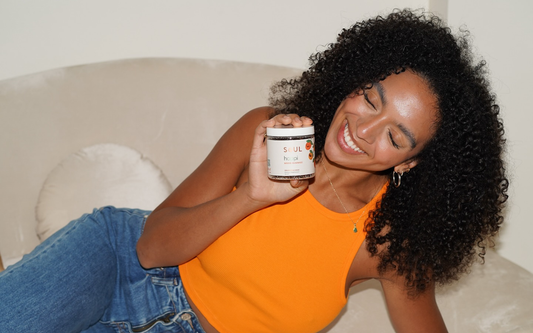Smiling woman wearing an orange tank top reclining on a beige couch holding a jar of Soul mood gummies