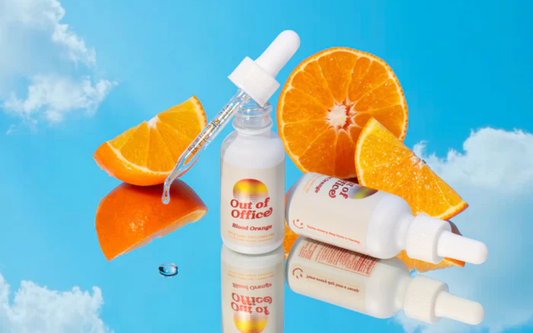 Three quarter view of Out of Office Blood Orange dropper bottle with droplet and sliced oranges on a reflective blue surface