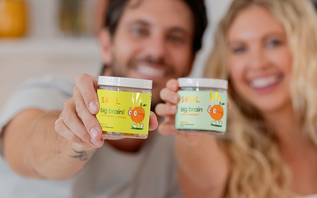 Two smiling people hold jars of Soul Big Brain focus gummies toward the camera in a bright home setting