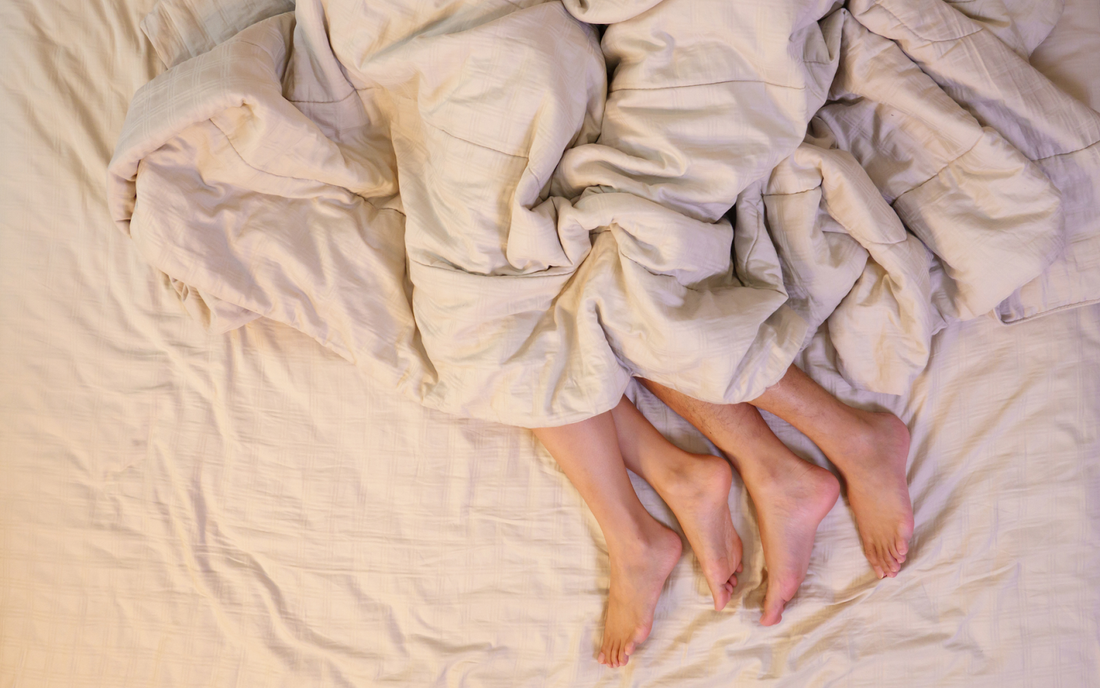 Overhead view of two people lying in bed with their legs entwined under a beige duvet