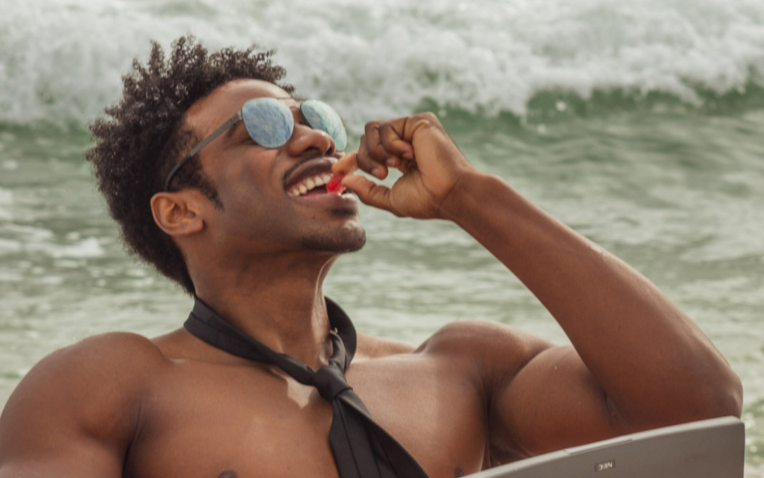 Shirtless man wearing sunglasses and a loosened black tie eating a red gummy by ocean waves