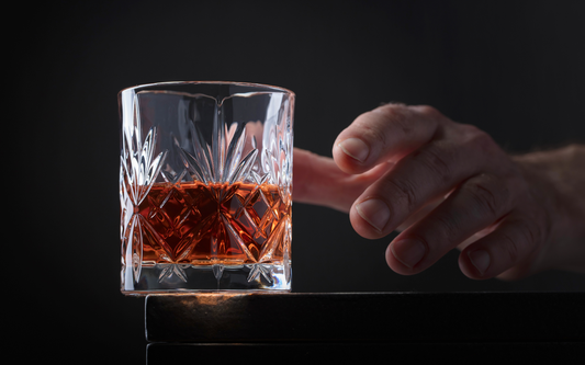 Crystal tumbler of amber whiskey with a hand reaching toward it on a dark background