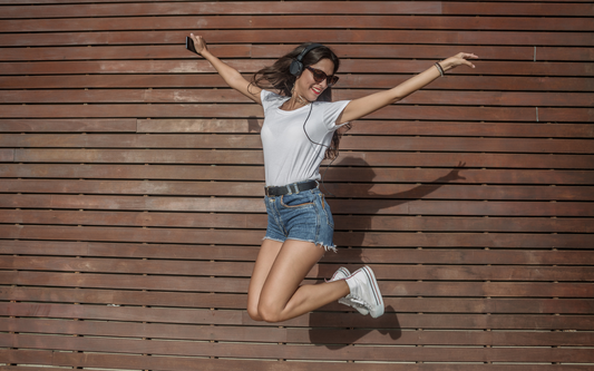 Woman jumping and smiling while wearing headphones and sunglasses in front of a wooden slat wall