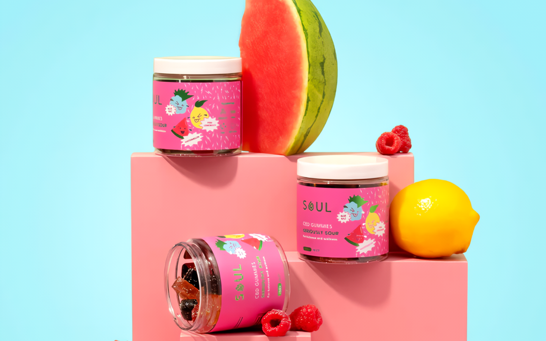 Front view of Soul CBD gummies jars with pink labels beside watermelon, raspberry, and lemon on pastel blue background