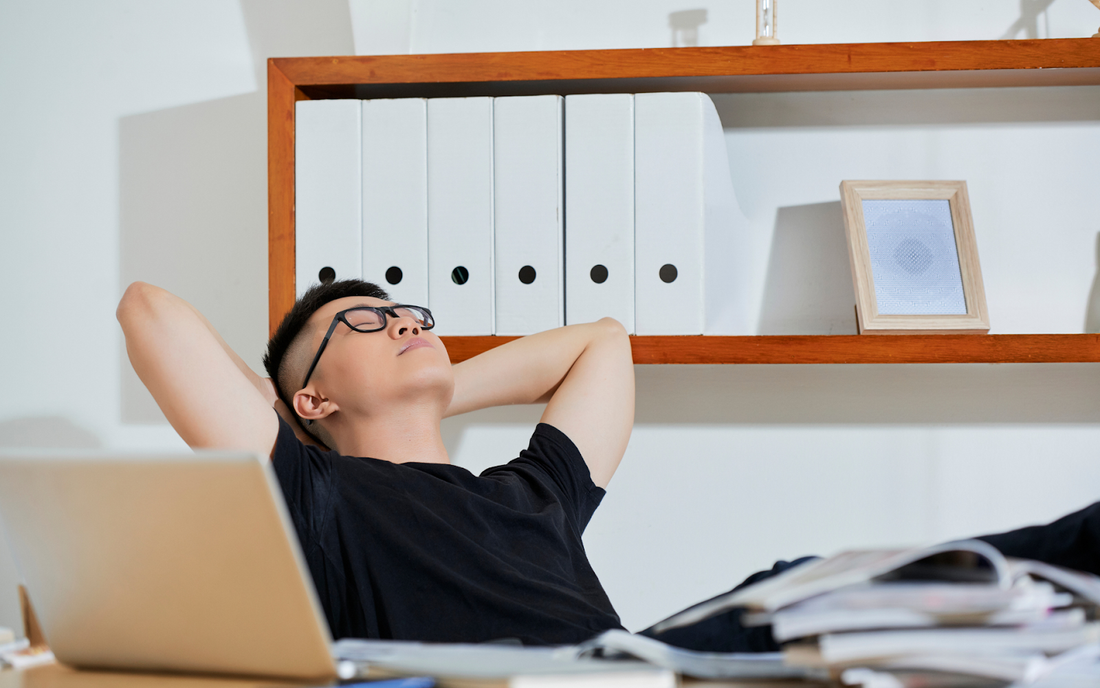 Young man reclining at a desk with laptop and stacked papers, hands behind his head and eyes closed, relaxing after work