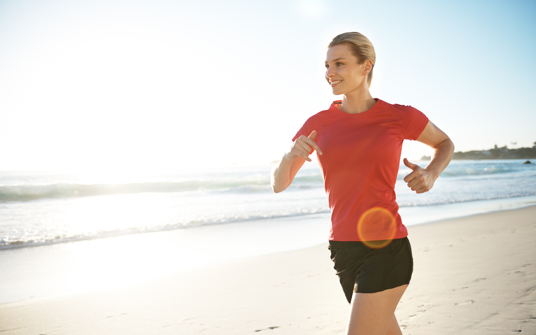 Woman jogging on a sunlit beach at sunrise, wearing a red shirt and black shorts